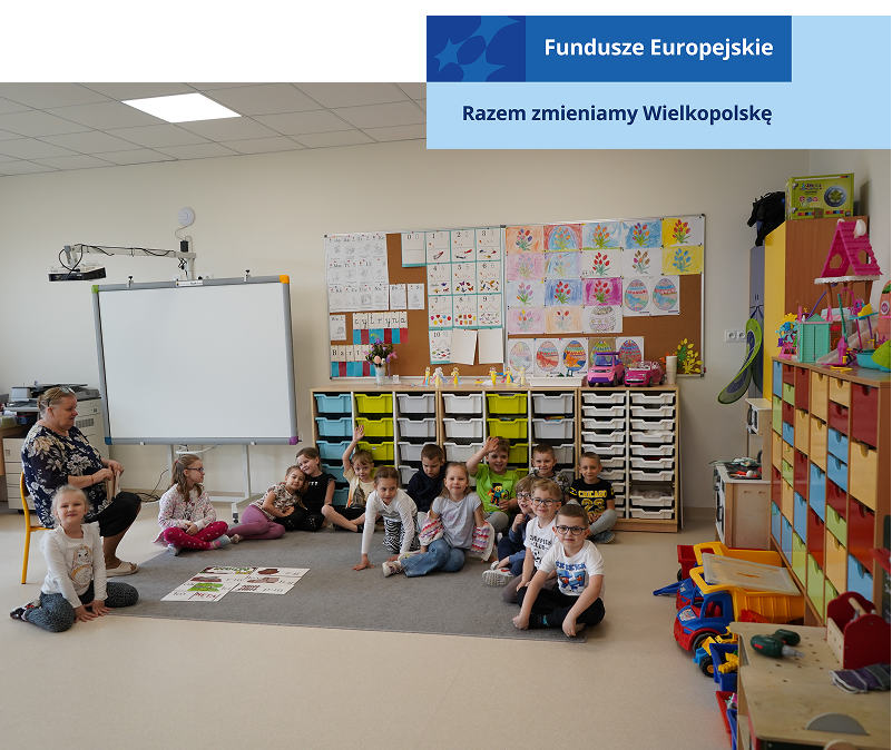 Fotografia przedstawia grupkę przedszkolaków siedzących na dywanie w sali. Obok, na krześle, siedzi nauczycielka. Autorem zdjęcia jest Sławomir Dawid. 