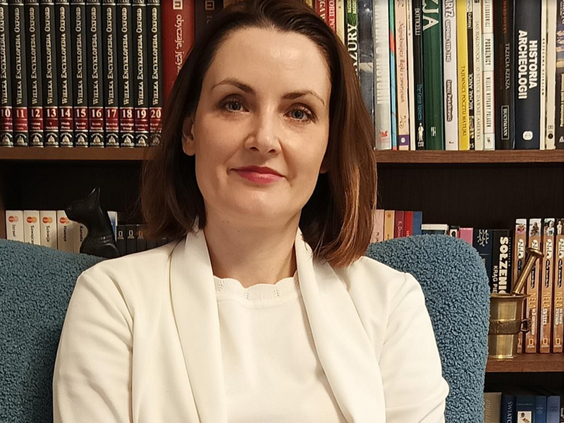 Zdjęcie przedstawia uśmiechniętą kobietę w średnim wieku, w eleganckim ubraniu. Siedzi w fotelu na tle książek. Fotografia pochodzi z archiwum prywatnego.