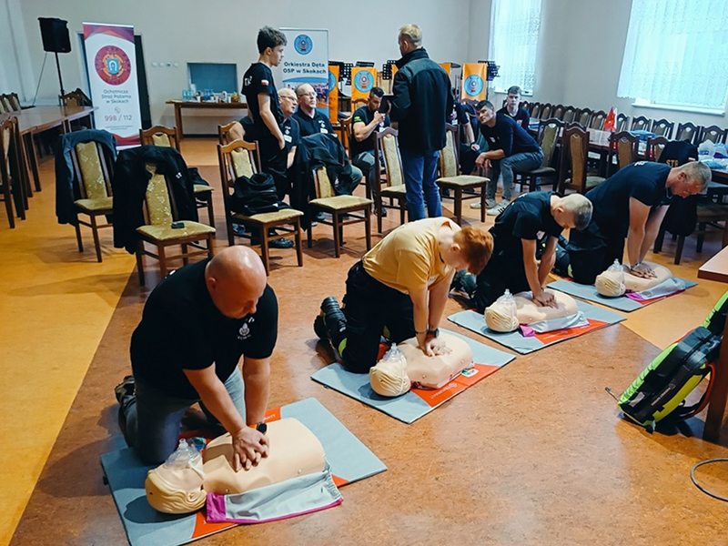Zdjęcie przedstawia szkolenie z pierwszej pomocy. Czterech mężczyzn wykonuje masaż serca na fantomie. Za nimi, na krzesłach, siedzi kilku mężczyzn, którzy się przyglądają. Fotografia pochodzi z archiwum Stowarzyszenia Ratujmy Życie.