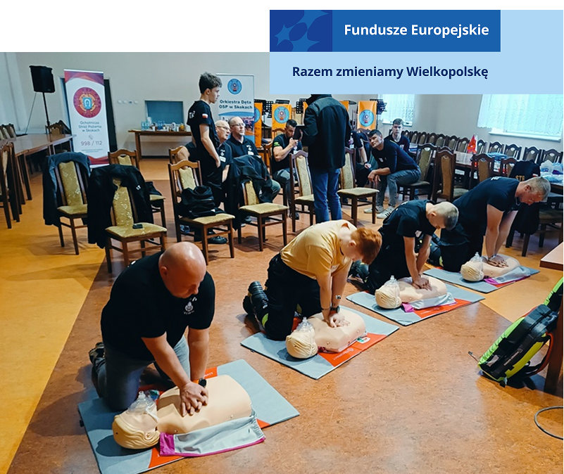 Zdjęcie przedstawia szkolenie z pierwszej pomocy. Czterech mężczyzn wykonuje masaż serca na fantomie. Za nimi, na krzesłach, siedzi kilku mężczyzn, którzy się przyglądają. Fotografia pochodzi z archiwum Stowarzyszenia Ratujmy Życie.