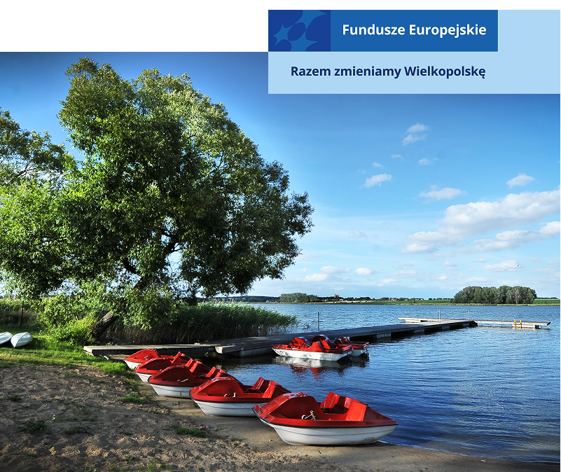 Na zdjęciu widoczne jest nabrzeże jeziora. Kilka czerwonych rowerków wodnych stoi na plaży, dwa kolejne zacumowane są do drewnianego pomostu. Dalej od zbiornika znajdują się obrócone do góry dnem, równo ustawione kajaki. Fotografia pochodzi z archiwum Stowarzyszenia Lokalnej Grupy Działania „Źródło”.