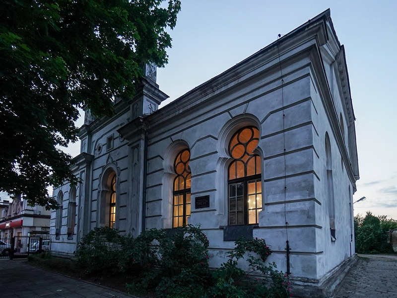 Zdjęcie przedstawia niewielki budynek dawnej synagogi, o jasnej elewacji. W środku świeci się światło. Fotografia pochodzi z archiwum beneficjenta.