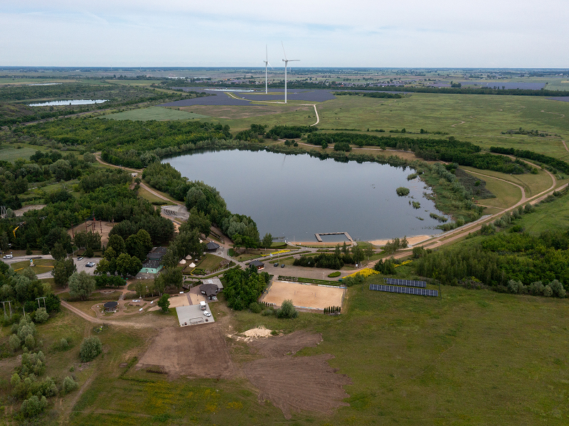 Zdjęcie z lotu ptaka przedstawia niewielkie jezioro otoczone lasami i polami. W jednym miejscu znajduje się infrastruktura rekreacyjna. W tle widoczne są dwa wiatraki. Fotografia pochodzi z archiwum beneficjenta.