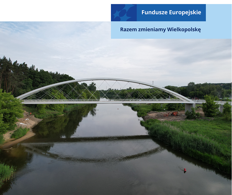 Zdjęcie przedstawia białą, łukową kładkę przerzuconą nad rzeką, łączącą dwa zielone brzegi. Fotografia pochodzi z archiwum Metropolii Poznań.