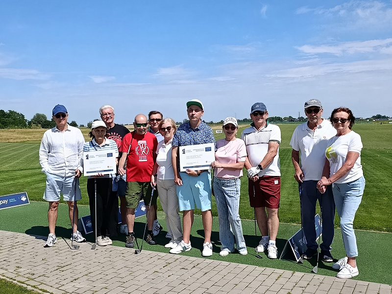 Na polu golfowym stoi grupa osób, głównie seniorzy. Niektórzy trzymają kije golfowe, inni tablice informacyjne dotyczące unijnego projektu. Autorem zdjęcia jest Łukasz Karkoszka.