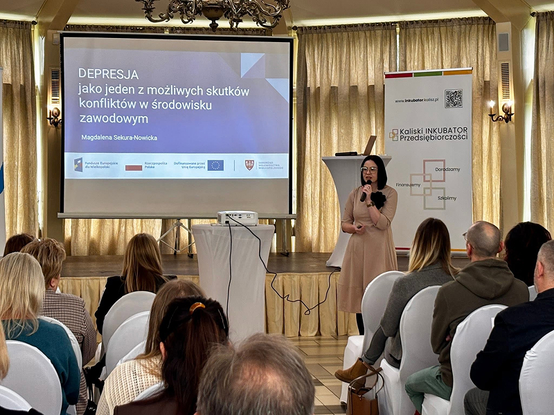 Zdjęcie wykonano w trakcie konferencji. Na scenie stoi kobieta, która przemawia do mikrofonu. Słucha jej licznie zebrana, siedząca publiczność. Fotografia pochodzi z archiwum beneficjenta.