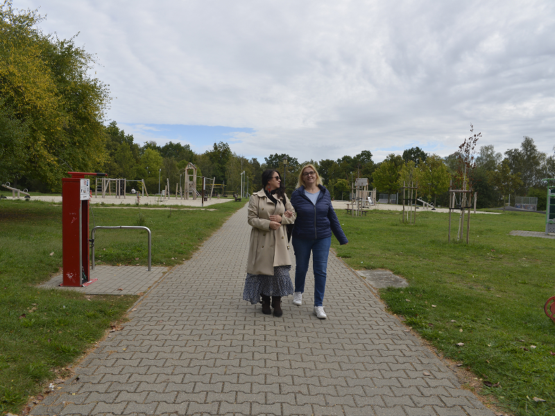 Zdjęcie przedstawia dwie panie, które spacerują parkową alejką, trzymając się pod rękę. W tle widoczna jest siłownia na świeżym powietrzu. Autorem fotografii jest Dominik Wójcik.
