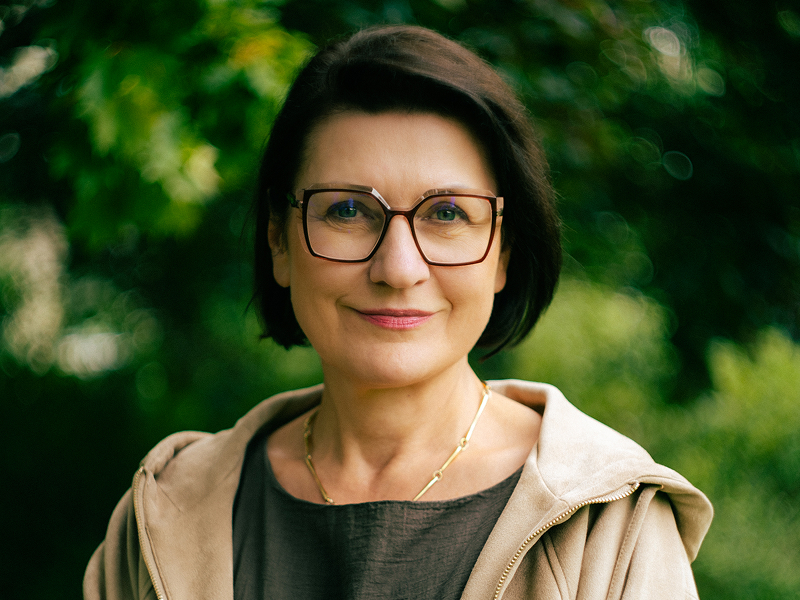 Zdjęcie przedstawia uśmiechniętą kobietę w średnim wieku, w okularach, na tle zieleni. Autorem fotografii jest Jacek Kordus.