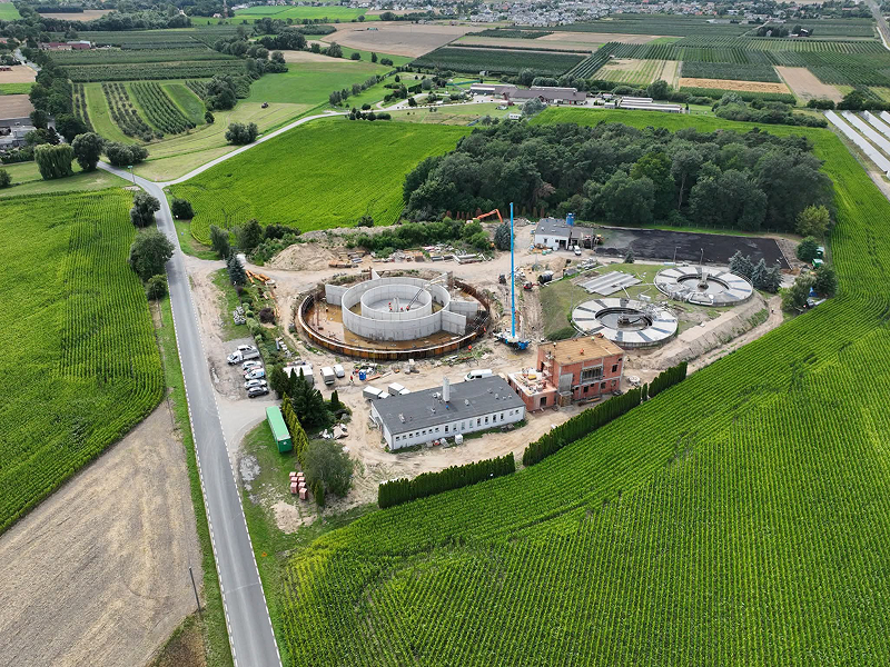 Zdjęcie z lotu ptaka przedstawia plac budowy oczyszczalni ścieków. Widoczne są wysokie dźwigi i powstające budynki. Fotografia pochodzi z archiwum beneficjenta.