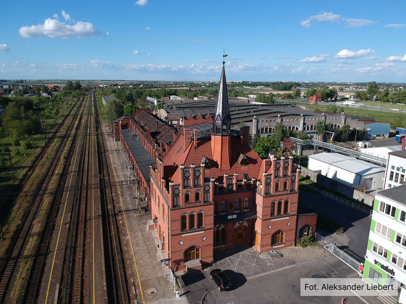 Fotografia z lotu ptaka przedstawia dworzec kolejowy, przypominający nieco w swojej budowie pałac – ma strzelistą wieżę, jest z czerwonej cegły. Z lewej znajdują się tory kolejowe, po prawej zaś zabudowania miejskie. Autorem zdjęcia jest Aleksander Liebert.