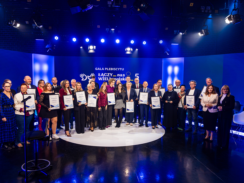 Na zdjęciu znajduje się grupa kilkudziesięciu osób, elegancko ubranych, które stoją na scenie i pozują do wspólnej fotografii. Scena ma miejsce w studio telewizyjnym, widoczne są więc niebieskie ekrany, kamery i oświetlenie. Zdjęcie pochodzi z archiwum Urzędu Marszałkowskiego Województwa Wielkopolskiego.