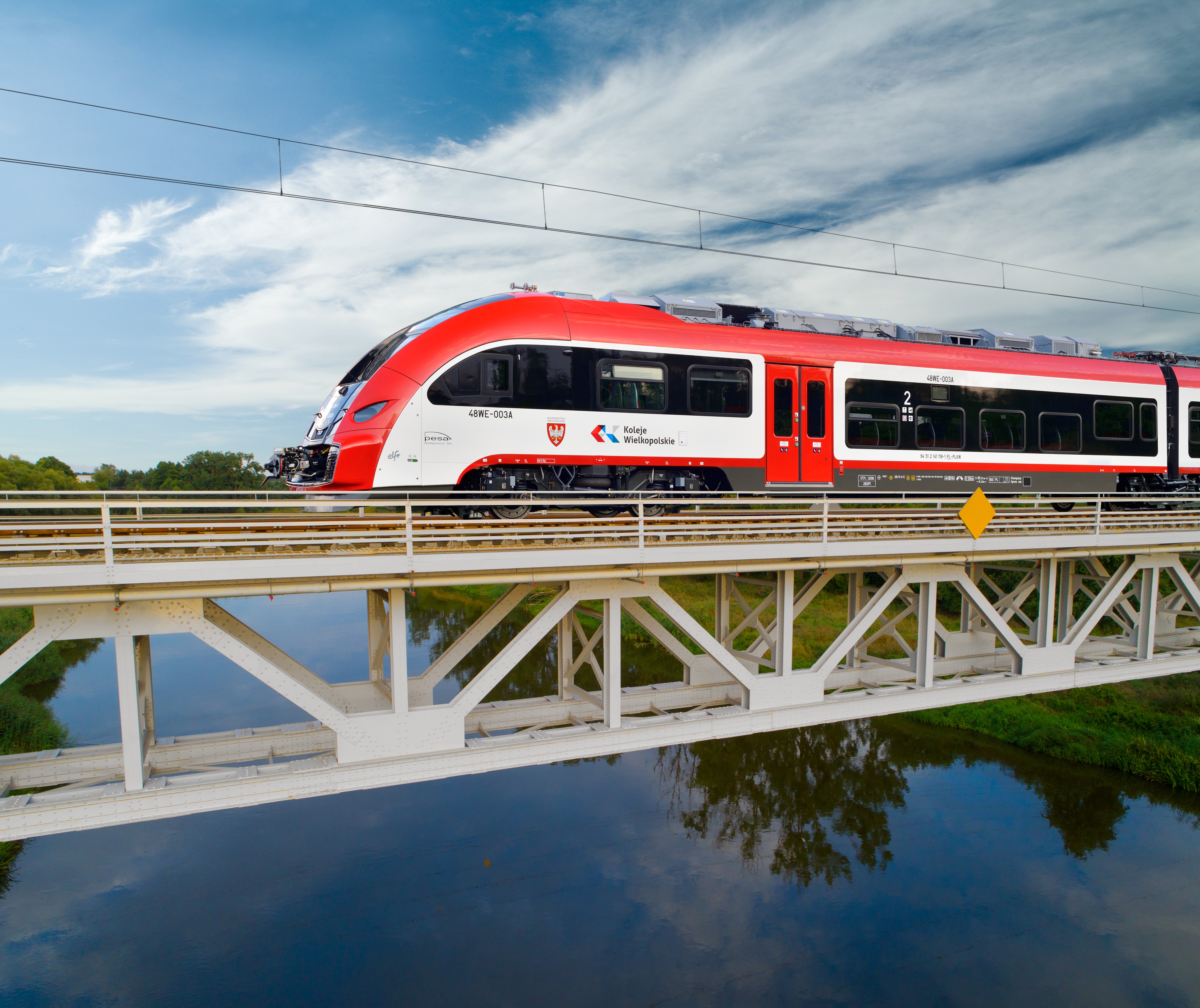 Na zdjęciu pociąg w barwach biało czerwonych, który przejeżdża przez most nad rzeką