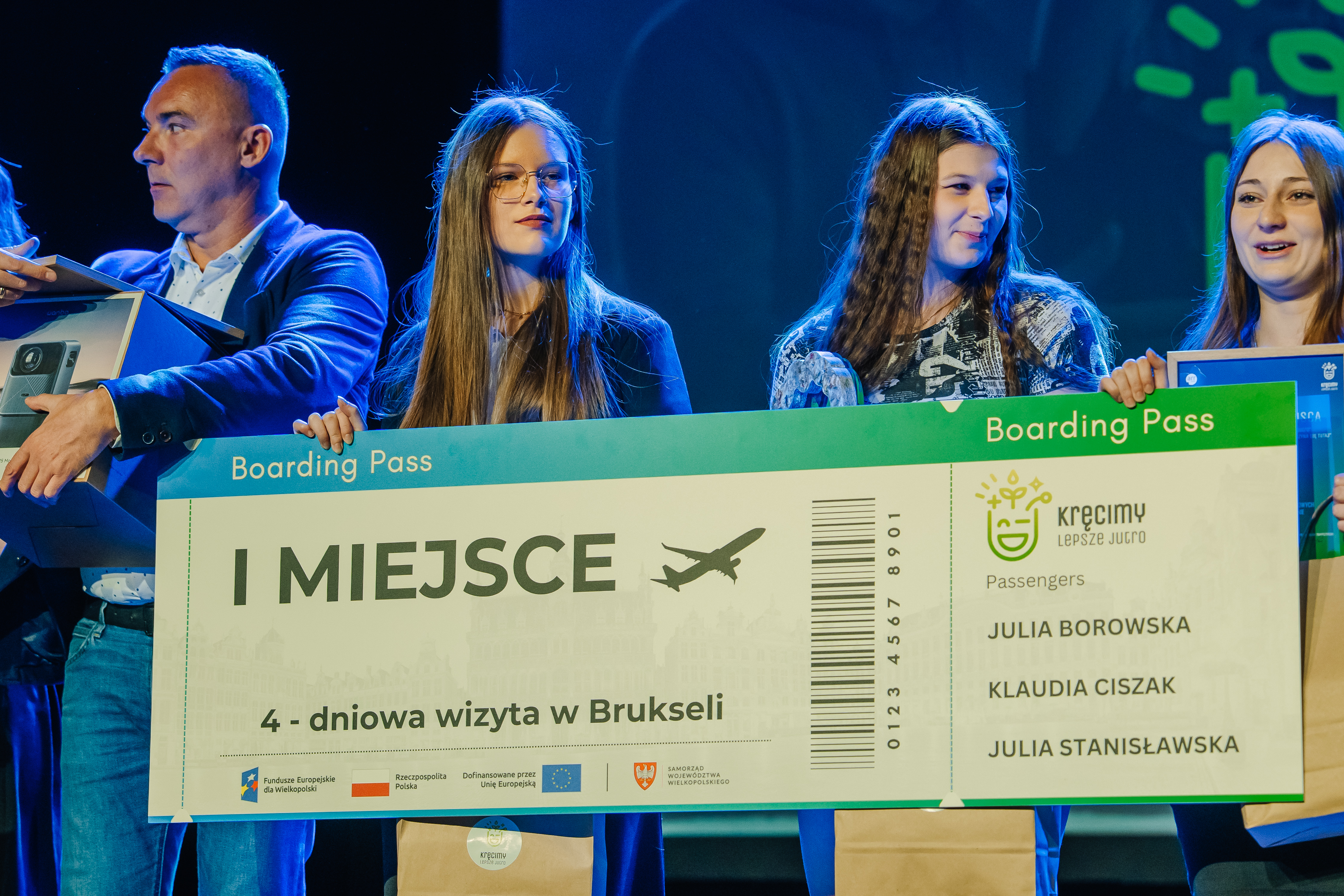 Trzy młode dziewczyny trzymające czek z napisem pierwsze miejsce. Z lewej strony stoi mężczyzna w marynarce