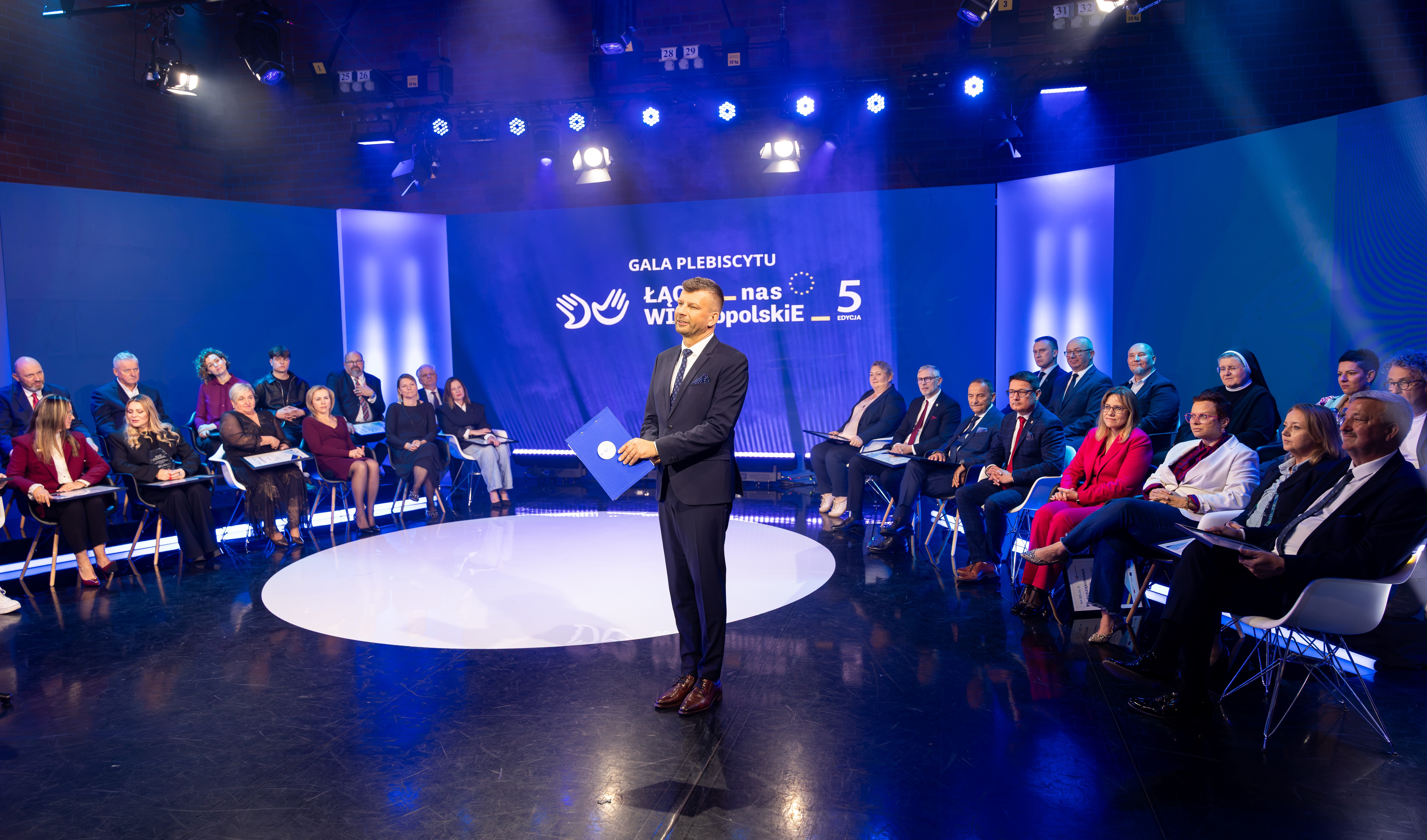 Na zdjęciu widać dużą scenę z osobami zasiadającymi na scenie, po środku mężczyzna w garniturze trzymający teczkę