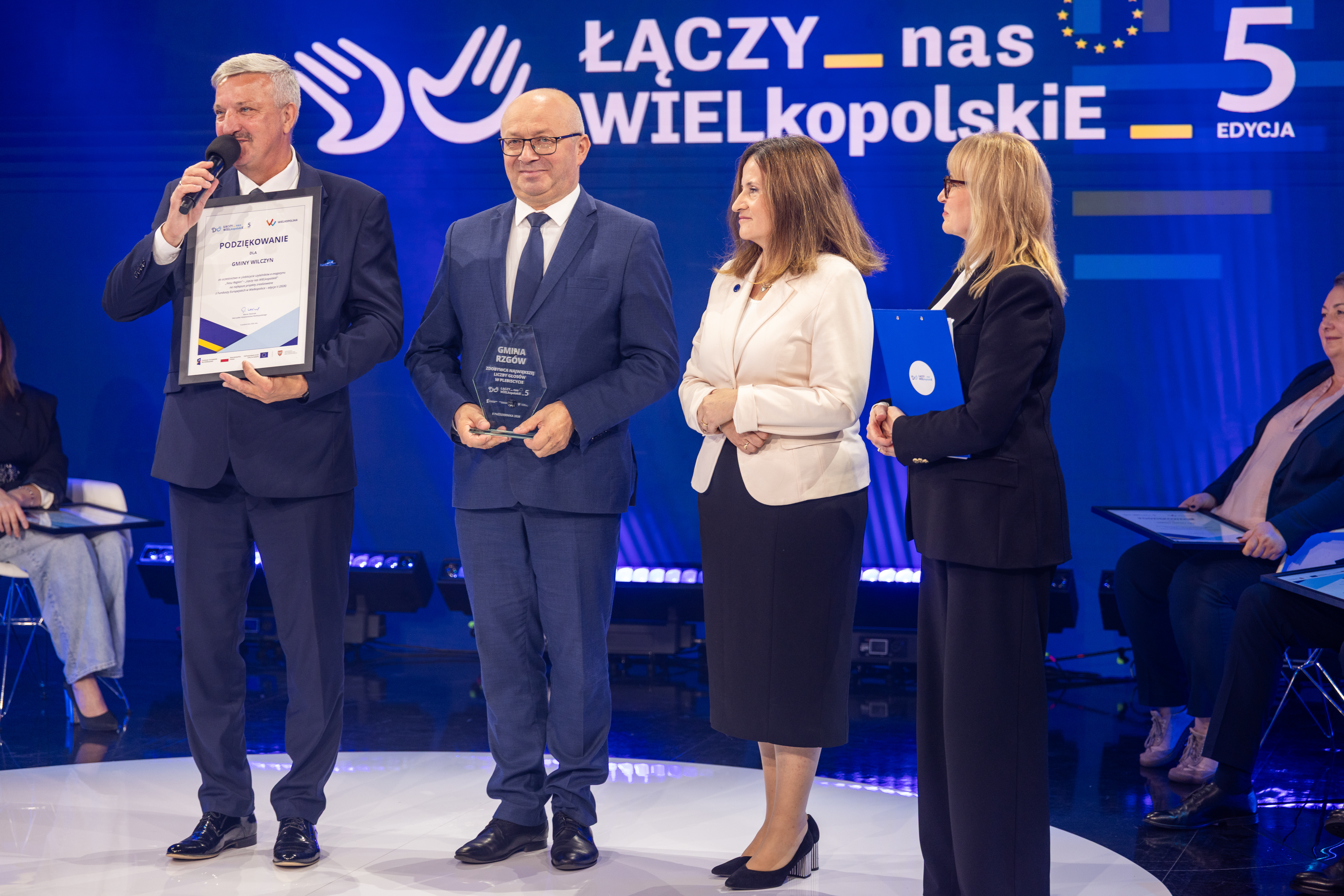 Na zdjęciu cztery osoby, jedna z nich przemawia do mikrofonu