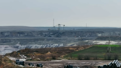 Wielka kopalnia odkrywkowa, z widoczną ogromną koparką wielonaczyniową w centrum, na tle rozległych hałd ziemi i węgla. Na pierwszym planie leżące rury i pola uprawne.