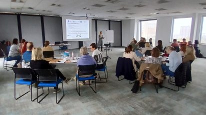 Sala konferencyjna wypełniona dorosłymi osobami siedzącymi na krzesłach i uczestniczącymi w prezentacji wyświetlanej na ekranie. Prowadząca stoi przy ekranie i tablicy.