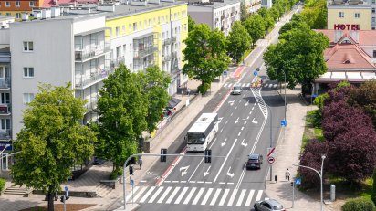 Widok z góry na wielopasmową miejską ulicę z przejściem dla pieszych, po której poruszają się samochody oraz biały autobus. Drogę otaczają zielone drzewa, wielopiętrowe bloki mieszkalne z jasnymi elewacjami oraz budynek z czerwonym dachem i napisem „HOTEL”.