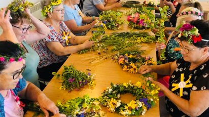 Grupa kobiet tworzy wianki z kwiatów, otoczona materiałami i narzędziami do rękodzieła.