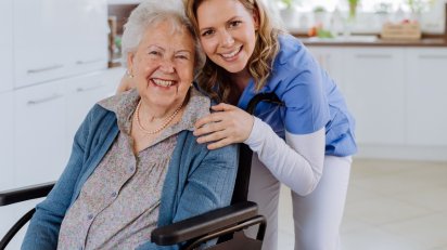 Starsza kobieta na wózku inwalidzkim przytula się z młodszą kobietą, obie uśmiechnięte, w przyjaznym otoczeniu.