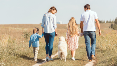 Rodzina z dwójką dzieci i białym psem spaceruje polną ścieżką wśród traw. Dorośli trzymają dzieci za ręce, wszyscy ubrani są swobodnie. W tle widać drzewa i jasne niebo.