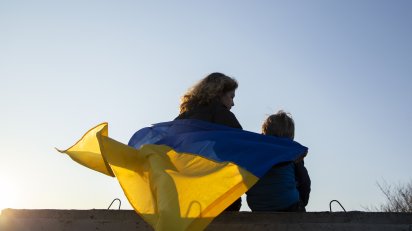 Kobieta i dziecko siedzą tyłem do zdjęcia na murze, okryci są ukraińską flagą.