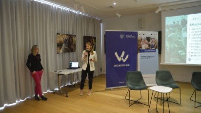 Dwie kobiety podczas prezentacji w sali konferencyjnej. Jedna stoi przy biurku i mówi do mikrofonu, a druga opiera się o ścianę. W tle widoczne są plakaty z logo "Wielkopolska" oraz ekran z wyświetlaną prezentacją.