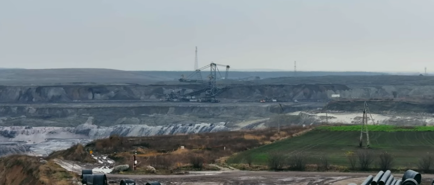 Wielka kopalnia odkrywkowa, z widoczną ogromną koparką wielonaczyniową w centrum, na tle rozległych hałd ziemi i węgla. Na pierwszym planie leżące rury i pola uprawne.