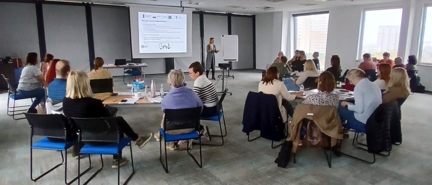 Sala konferencyjna wypełniona dorosłymi osobami siedzącymi na krzesłach i uczestniczącymi w prezentacji wyświetlanej na ekranie. Prowadząca stoi przy ekranie i tablicy.