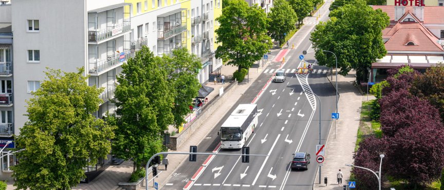 Widok z góry na wielopasmową miejską ulicę z przejściem dla pieszych, po której poruszają się samochody oraz biały autobus. Drogę otaczają zielone drzewa, wielopiętrowe bloki mieszkalne z jasnymi elewacjami oraz budynek z czerwonym dachem i napisem „HOTEL”.