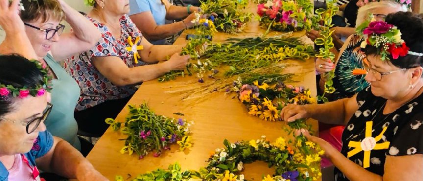 Grupa kobiet tworzy wianki z kwiatów, otoczona materiałami i narzędziami do rękodzieła.