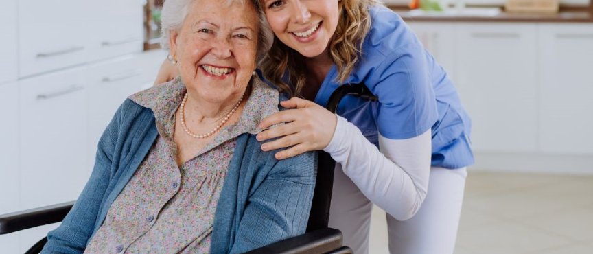 Starsza kobieta na wózku inwalidzkim przytula się z młodszą kobietą, obie uśmiechnięte, w przyjaznym otoczeniu.