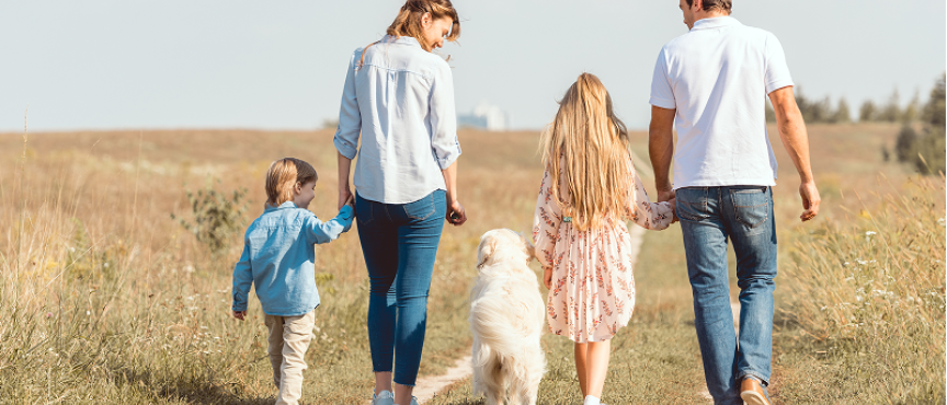 Rodzina z dwójką dzieci i białym psem spaceruje polną ścieżką wśród traw. Dorośli trzymają dzieci za ręce, wszyscy ubrani są swobodnie. W tle widać drzewa i jasne niebo.