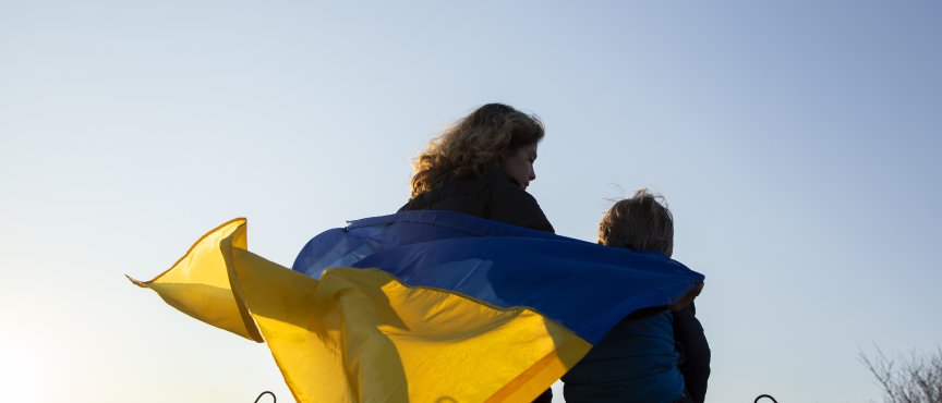 Kobieta i dziecko siedzą tyłem do zdjęcia na murze, okryci są ukraińską flagą.