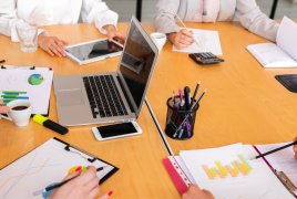 grupa osób przy stole, na którym widać rozłożone laptopy, tablety i notatniki