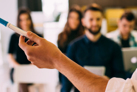 grupa osób siedzących w sali, na pierwszym planie widać uniesioną rękę z długopisem w dłoni