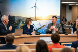 Spotkanie na sali, na zdjęciu widać ludzi na widowni oraz stół prezydialny z siedzącymi przy nim osobami, w tle zdjęcie szybu kopalni i wiatraków