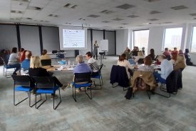 Sala konferencyjna wypełniona dorosłymi osobami siedzącymi na krzesłach i uczestniczącymi w prezentacji wyświetlanej na ekranie. Prowadząca stoi przy ekranie i tablicy.