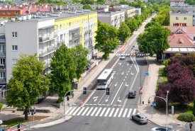 Widok z góry na wielopasmową miejską ulicę z przejściem dla pieszych, po której poruszają się samochody oraz biały autobus. Drogę otaczają zielone drzewa, wielopiętrowe bloki mieszkalne z jasnymi elewacjami oraz budynek z czerwonym dachem i napisem „HOTEL”.