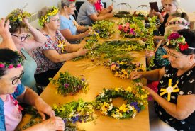 Grupa kobiet tworzy wianki z kwiatów, otoczona materiałami i narzędziami do rękodzieła.