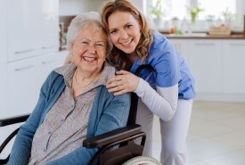 Starsza kobieta na wózku inwalidzkim przytula się z młodszą kobietą, obie uśmiechnięte, w przyjaznym otoczeniu.