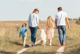 Rodzina z dwójką dzieci i białym psem spaceruje polną ścieżką wśród traw. Dorośli trzymają dzieci za ręce, wszyscy ubrani są swobodnie. W tle widać drzewa i jasne niebo.
