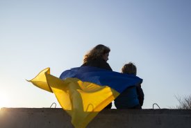 Kobieta i dziecko siedzą tyłem do zdjęcia na murze, okryci są ukraińską flagą.