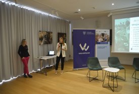 Dwie kobiety podczas prezentacji w sali konferencyjnej. Jedna stoi przy biurku i mówi do mikrofonu, a druga opiera się o ścianę. W tle widoczne są plakaty z logo "Wielkopolska" oraz ekran z wyświetlaną prezentacją.