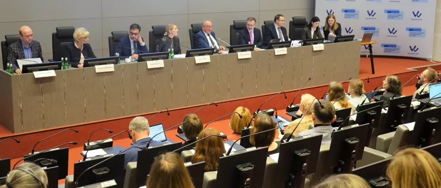 Na zdjęciu widać stół prezydialny z ośmioma osobami i kilknaście osób siedzący po drugiej stronie