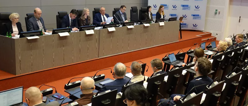 Na zdjęciu widać osiem osób zasiadających przy stole prezydialnym i kilkanaście osób siedzący na widowni. W tle ścianka Funduszy Europejskich