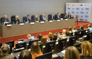 Na zdjęciu widać stół prezydialny z ośmioma osobami i kilknaście osób siedzący po drugiej stronie