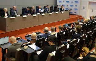 Na zdjęciu widać osiem osób zasiadających przy stole prezydialnym i kilkanaście osób siedzący na widowni. W tle ścianka Funduszy Europejskich