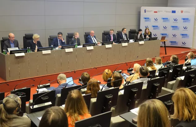 Na zdjęciu widać stół prezydialny z ośmioma osobami i kilknaście osób siedzący po drugiej stronie