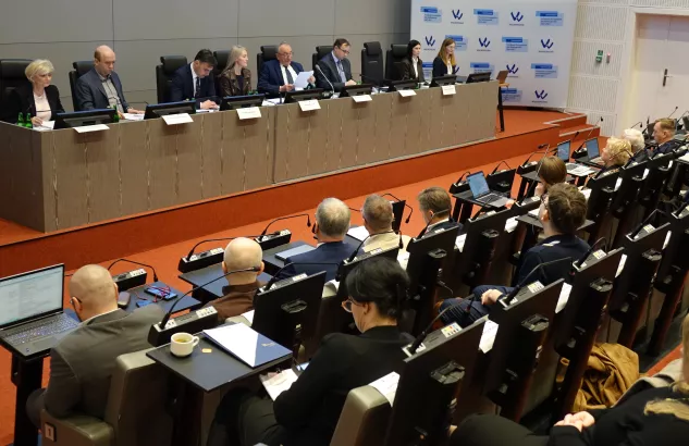 Na zdjęciu widać osiem osób zasiadających przy stole prezydialnym i kilkanaście osób siedzący na widowni. W tle ścianka Funduszy Europejskich